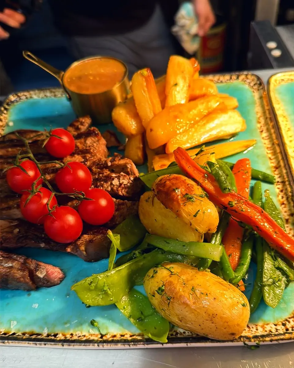 A grill platter with tomatoes, chips and vegetables on a turquoise board at the pass
